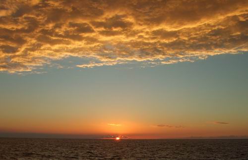 Golden clouds stretch across the sky over a calm ocean at sunset, with the sun glowing orange near the horizon and reflecting on the water.
