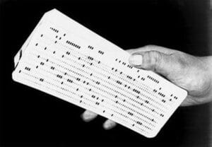 A hand holds a stack of punched cards, used for data storage and programming in early computers. The cards have rows of rectangular holes punched in patterns.