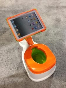 A child’s potty training seat with an attached tablet holder displaying a tablet, placed on a gray carpeted floor. The potty is white with orange and green accents.