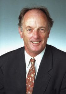 A middle-aged man with thinning hair, wearing a dark suit, white shirt, and patterned tie, smiling at the camera against a plain, light-colored background.