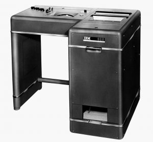 Black and white photo of an IBM Test Scoring machine with a compact rectangular design, control knobs, and slots for inserting and processing tests.