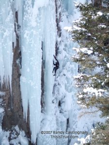 A person wearing winter gear climbs a vertical pillar of ice on a cliffside using ice axes, with snow-covered trees nearby.