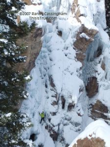 A person in a bright yellow jacket climbs a snowy, rocky cliff covered in ice and icicles, surrounded by trees.