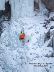 A person wearing a bright orange jacket and yellow helmet is ice climbing a steep, snow-covered frozen waterfall with ice axes, surrounded by rocky cliffs.