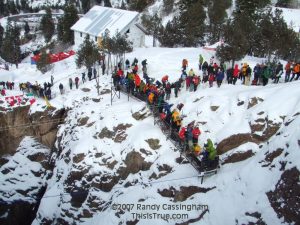 A crowd in winter clothing gathers on a snowy cliff and a narrow bridge spanning a deep gorge. Snow covers the ground, and pine trees and buildings are visible in the background.