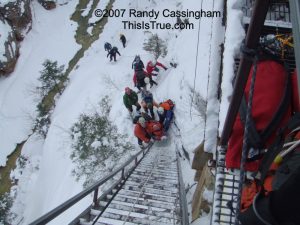 A group of people, wearing winter gear, carrying an injured climber in a rescue basket, climb a steep metal ladder up a snowy cliffside while others wait below. Snow and trees cover the landscape, and gear is visible on the metal platform above.