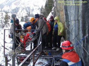 A group of rescue workers in winter gear assist a person on a stretcher on a snowy mountain ledge, using ropes and safety equipment next to a rocky cliff. Trees and mountains are visible in the background.