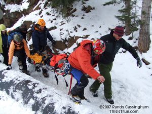 A group of rescuers in winter gear carry an injured person on a stretcher up a snowy slope in a forested area, working together to navigate the icy terrain.