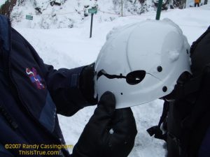 A person holds a white helmet with a large, jagged crack down the middle, showing snow on it. The background is snowy, and the person wears a blue jacket with an EMS logo.