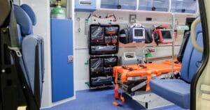 Interior of an ambulance showing medical equipment, an orange stretcher with straps, blue padded seats, oxygen tanks, and emergency bags organized along the wall.