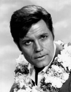 Black and white portrait of actor Jack Lord, with dark hair wearing a suit and a floral lei around his neck, looking slightly past the camera with a neutral expression.