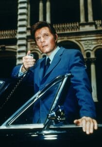 Jack Lord in a blue suit standing beside an open car door, holding a radio microphone and looking intently ahead, with a historic building featuring arches in the background.