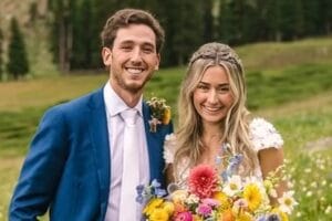 A smiling couple poses outdoors on their wedding day; the groom wears a blue suit and tie, and the bride wears a white dress and holds a colorful bouquet, with greenery and trees in the background.