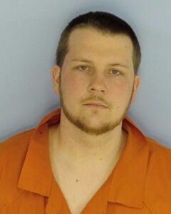 Mug shot of a young adult man with short brown hair and a trimmed beard wears an orange prison jumpsuit and faces the camera with a neutral expression against a plain light blue background.