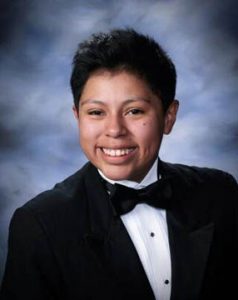 A young woman with short dark hair, dressed in a black tuxedo, white shirt, and black bow tie, smiling in front of a cloudy blue and gray studio backdrop.