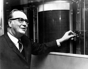 Rey Johnson in a suit and glasses stands next to an early computer hard drive. It has multiple platters that can be seen through a glass window. He smiling and touching what appears to be the device that moves the read/write heads between the platters.