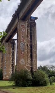 A tall abandoned railroad bridge seen from the ground below. The pillars are heavily tagged with graffiti.