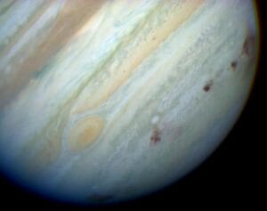 A close-up image of Jupiter shows its swirling cloud bands, the Great Red Spot on the left, and several dark marks from the Shoemaker-Levy 9 comet impacts on the planet's surface.