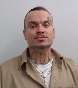 A man with short hair, facial hair, and multiple tattoos on his face and neck wears a tan collared shirt over a white undershirt and several silver necklaces, standing against a plain light-colored background.
