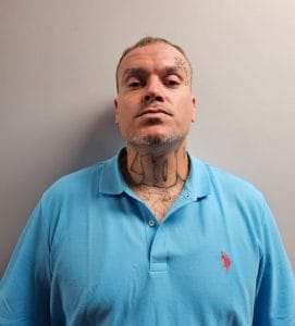 A man in a light blue polo shirt stands against a plain wall. He has short hair, visible tattoos on his neck and face, and a neutral facial expression.