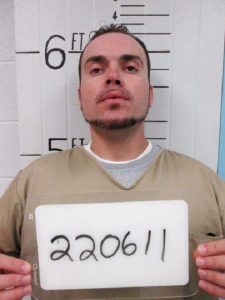 A man in a tan prison uniform stands against a height chart holding a sign with the number 220611 written on it. The chart shows he is 6 feet, 2 inches tall.