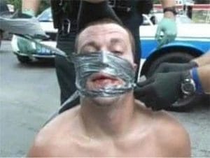 A man appears to be kneeling on the ground, not wearing a shirt, with his face partially wrapped in duct tape. A police officer with gloved hands is unwrapping the tape.