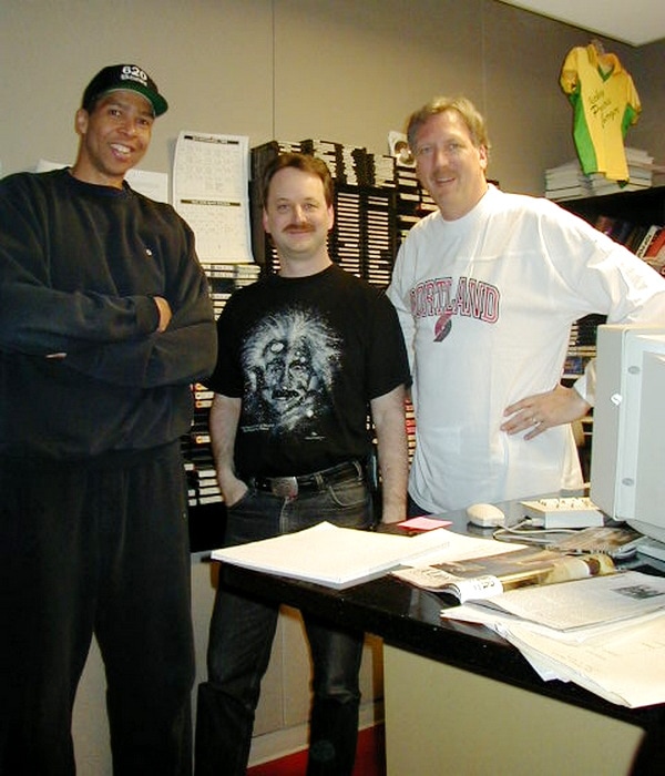 Three men standing and smiling in an office. One wears a black cap and sweatshirt, the second wears a black shirt with Einstein’s face, and the third wears a white “Portland” shirt. Papers and office equipment are on the desk. Randy, in the middle, is clearly much shorter than the other two men.