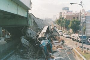 A collapsed elevated highway with a large truck crushed beneath the debris. Emergency vehicles and people are present at the scene, and damaged buildings are visible in the background.