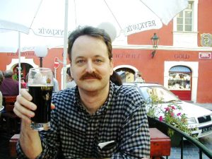 Me in a checkered shirt, looking into the camera while sitting at a table under an umbrella in Prague, Czech Republic, holding a partly-consumed half-liter of dark beer.