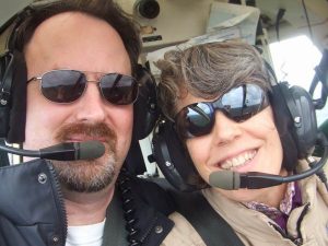Randy and Kit wearing headsets and sunglasses smile inside an aircraft cockpit, with controls and windows visible in the background.