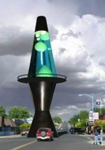 A giant lava lamp sculpture in the middle of a suburban intersection. It towers over the street with cars and people, with an observation deck half-way up, resembling an actual lava lamp with green blobs inside, set against a cloudy sky.