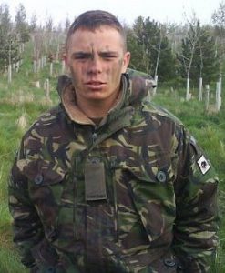 A man in camouflage military attire stands on grassy ground outdoors, with face paint applied and trees visible behind him.