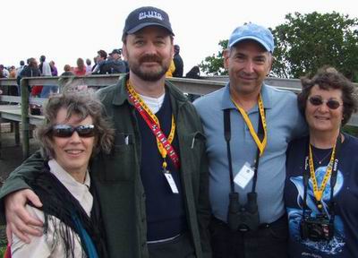 Four adults wearing access badges smile for a group photo outdoors. Two men stand in the middle, both wearing caps. From left to right is Kit Cassingham, Randy Cassingham, David Levy, and Wendee Levy. Other people are in the background.