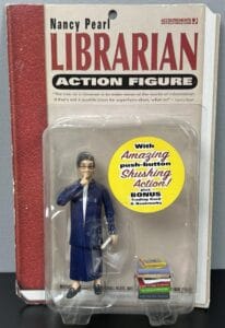 A Nancy Pearl Librarian Action Figure in packaging, featuring a doll in a blue outfit with glasses, a push-button shushing action, and small book accessories. The packaging resembles a red and white book cover.