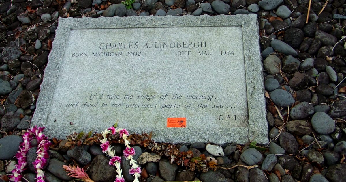 A gravestone for Charles A. Lindbergh, surrounded by flowers, lies on dark rocks. The inscription notes his birth in Michigan in 1902 and death in Maui in 1974, along with a poetic quote at the bottom. An orange card can be seen on the marker.