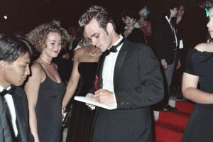 A young man in a tuxedo signs an autograph while standing on a red carpet, surrounded by people dressed in formal evening wear.