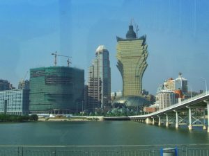 A city skyline with modern high-rise buildings, including a uniquely shaped, gold-colored tower; a construction site and crane are visible. A body of water and a white bridge are in the foreground under a clear blue sky.