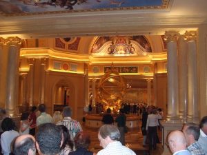 A crowd of people stands inside a grand, ornate lobby with tall columns, golden decor, and a large golden armillary sphere statue in the center. The ceiling is elaborately painted with classical artwork.