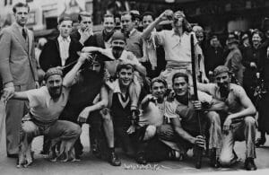 A group of men in costume, some dressed as pirates, pose energetically together on a city street, while a small crowd of onlookers stands in the background, watching the scene.