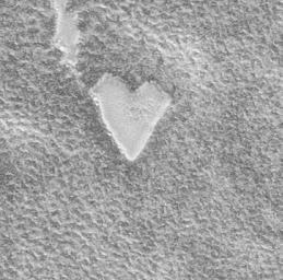A heart-shaped patch of lighter texture stands out against a rough, grainy gray surface, resembling frost or snow. It's a photo from Mars.