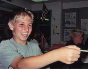 A smiling boy in a gray T-shirt holds a pencil in a classroom setting, with other students and educational posters visible in the background.
