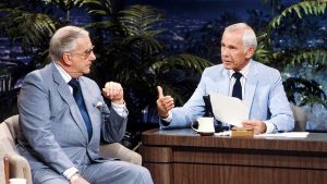 Two men in suits sit and talk on a late-night talk show set. One is behind a desk holding papers, and the other sits in a guest chair. They appear engaged in conversation, with plants and city lights in the background.