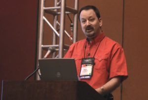 Randy Cassingham in a red shirt stands at a lectern with a laptop and microphone, speaking in front of a brown wall and metal truss structure. He is wearing a conference badge and gesturing with his right hand.