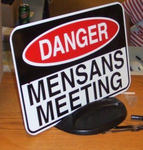 A sign on a desk reads DANGER MENSANS MEETING in bold letters, with the word DANGER in white text on a red oval, resembling a caution or warning sign.