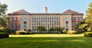 A large brick and stone academic building with symmetrical windows, a central entrance, and a small cupola on the roof, surrounded by green lawns and trees on a sunny day.