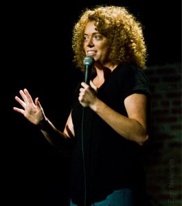 Comedian Michelle Wolf on stage performing. She is dressed in black against a black background, and is speaking into a microphone as she gestures with her other hand.