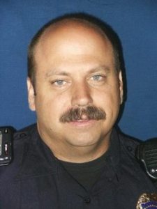 An official portrait of a middle-aged man with light skin, short hair, and a mustache wearing a dark police uniform stands in front of a plain blue background. He has a radio mic clipped to his shoulder.