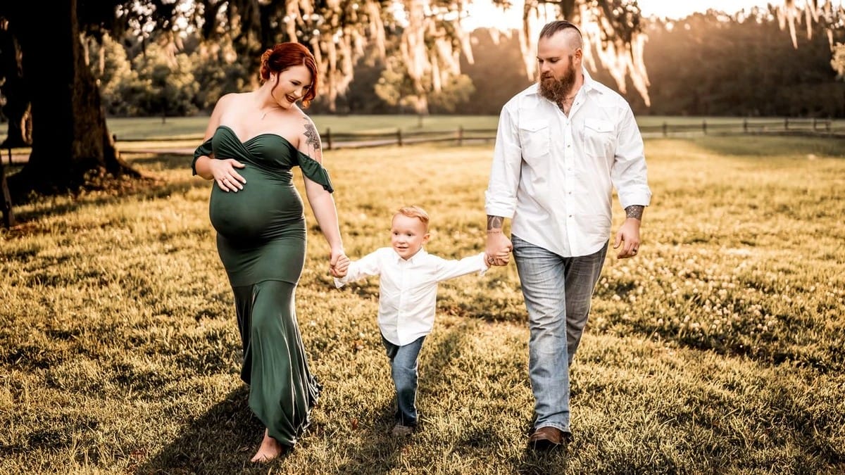 A pregnant woman in a green dress walks hand-in-hand with a bearded man and a young boy across a sunlit grassy field, all smiling and enjoying the outdoors together.