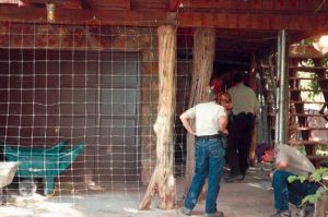Two wildlife officers in uniforms examine the ground near a fenced area outside a rustic home, while two others stand near a doorway and stairs. A green wheelbarrow is visible on the left: it held dog food.