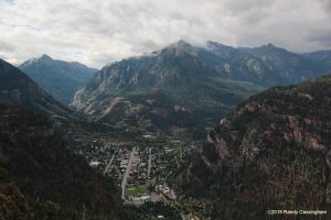 Ouray, Colorado: a tiny town nestled in a tight valley surrounded by tall, forested mountains under a cloudy sky, with roads and buildings visible below.
